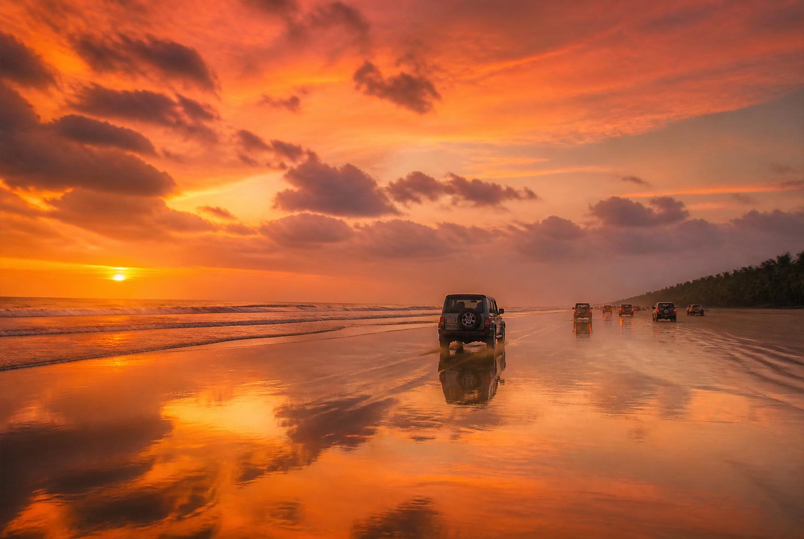 Cars driving along Muzhappilangad Drive-in Beach at sunset with orange sky reflections