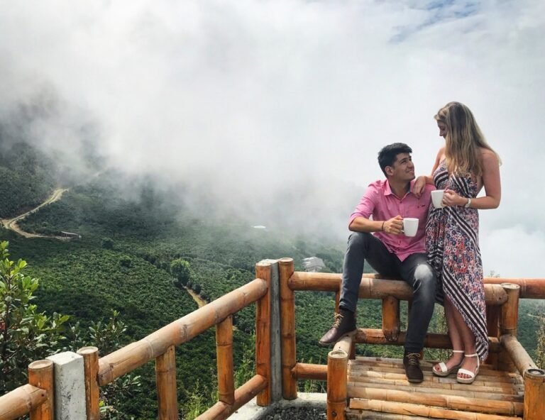 A couple sitting on a wooden railing amid misty green hills, holding warm drinks
