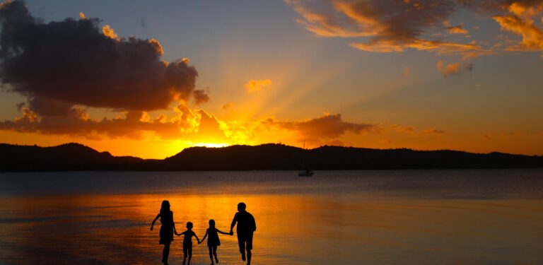 A family of four silhouetted against a golden sunset beside calm water